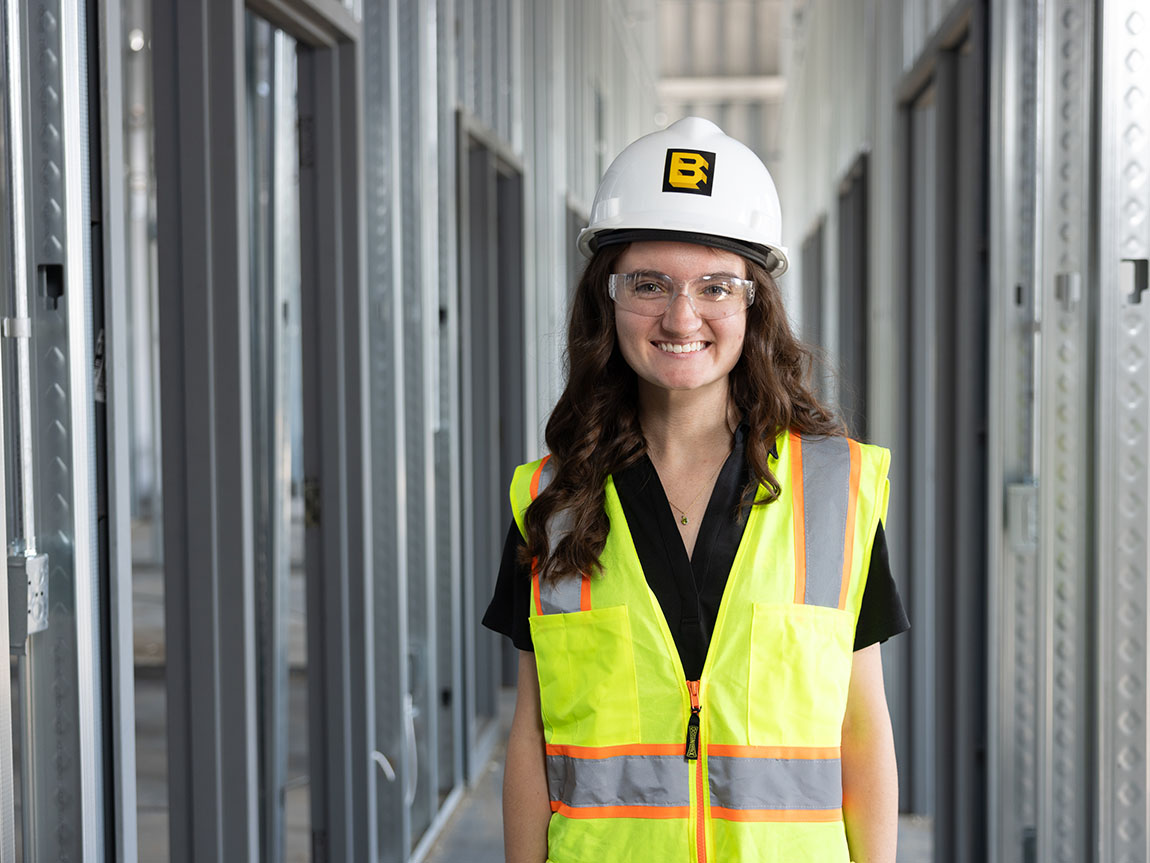 A young adult in a construction helmet and vest smiles for the camera